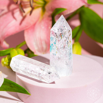 The Limitless Manifestation - Ice Quartz Point by Conscious Items features two crystal points on a light pink stand, accented by a pink lily and green leaves. Bright lighting highlights their clarity, making them powerful manifestation tools for any space.