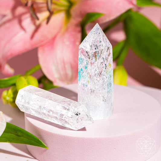 The Limitless Manifestation - Ice Quartz Point by Conscious Items features two crystal points on a light pink stand, accented by a pink lily and green leaves. Bright lighting highlights their clarity, making them powerful manifestation tools for any space.