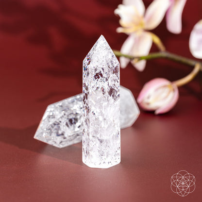 The Limitless Manifestation - Ice Quartz Point by Conscious Items stands upright on a red surface beside another, with soft pink orchids in the background. This crystal is a powerful amplifier, channeling omni-directional manifestation energy.