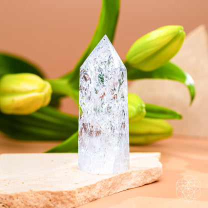 The Limitless Manifestation - Ice Quartz Point by Conscious Items stands upright on a light stone slab to amplify your intentions. Green lily buds and leaves blur against a peach backdrop, with a white geometric pattern at the bottom right.