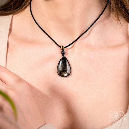 Close-up of a person wearing Conscious Items’ Hypersthene Millionaire Manifestation Pendant—a black cord necklace with a teardrop-shaped Hypersthene stone—against bare skin and a beige top, hand resting near the collarbone.