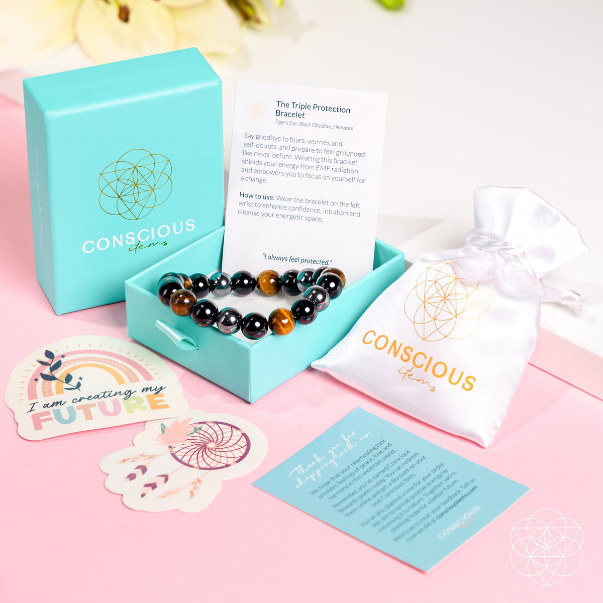 An open turquoise box from Conscious Items displays The Triple Protection Bracelet, protective stones, an info card, white pouch, and motivational cards (“I am creating my future,” “I always feel protected”) on a white surface with yellow flowers.