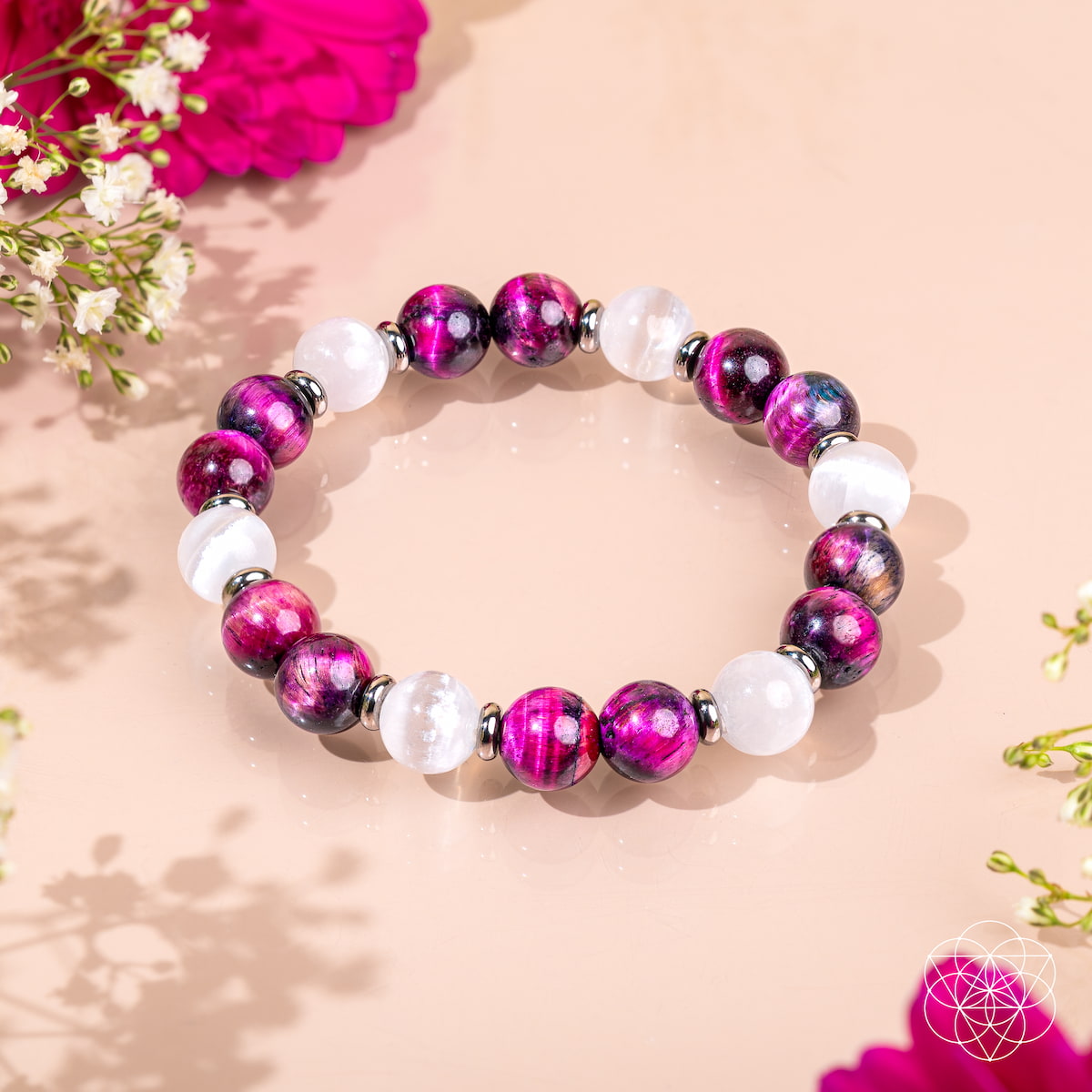 The Shape Your Future – Pink Galaxy Tiger’s Eye Bracelet by Conscious Items features alternating glossy purple and white beads with silver spacers, displayed in a circle on a light pink surface with delicate flowers and petals.