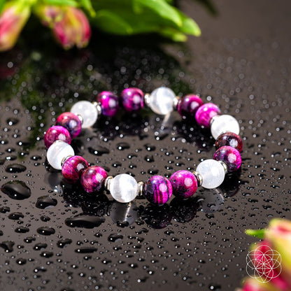 The Shape Your Future – Pink Galaxy Tiger’s Eye Bracelet by Conscious Items features alternating purple and white stones on a wet black surface, with water droplets and blurred green leaves and pink flowers in the background.