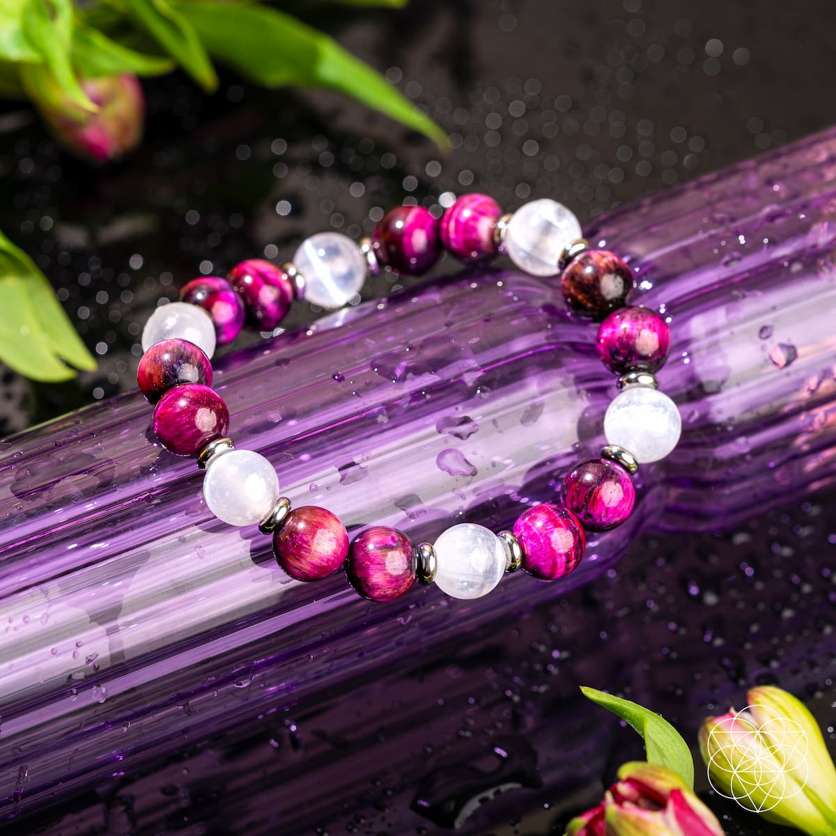 The Shape Your Future – Pink Galaxy Tiger’s Eye Bracelet by Conscious Items features alternating pink and white stones with metallic spacers, displayed on a wet purple surface with green leaves in the background.