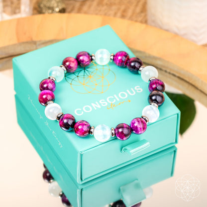 The Shape Your Future – Pink Galaxy Tiger’s Eye Bracelet by Conscious Items, featuring purple, magenta, and white beads, is displayed on a turquoise Conscious Items box atop a mirrored surface beside manifestation crystals.