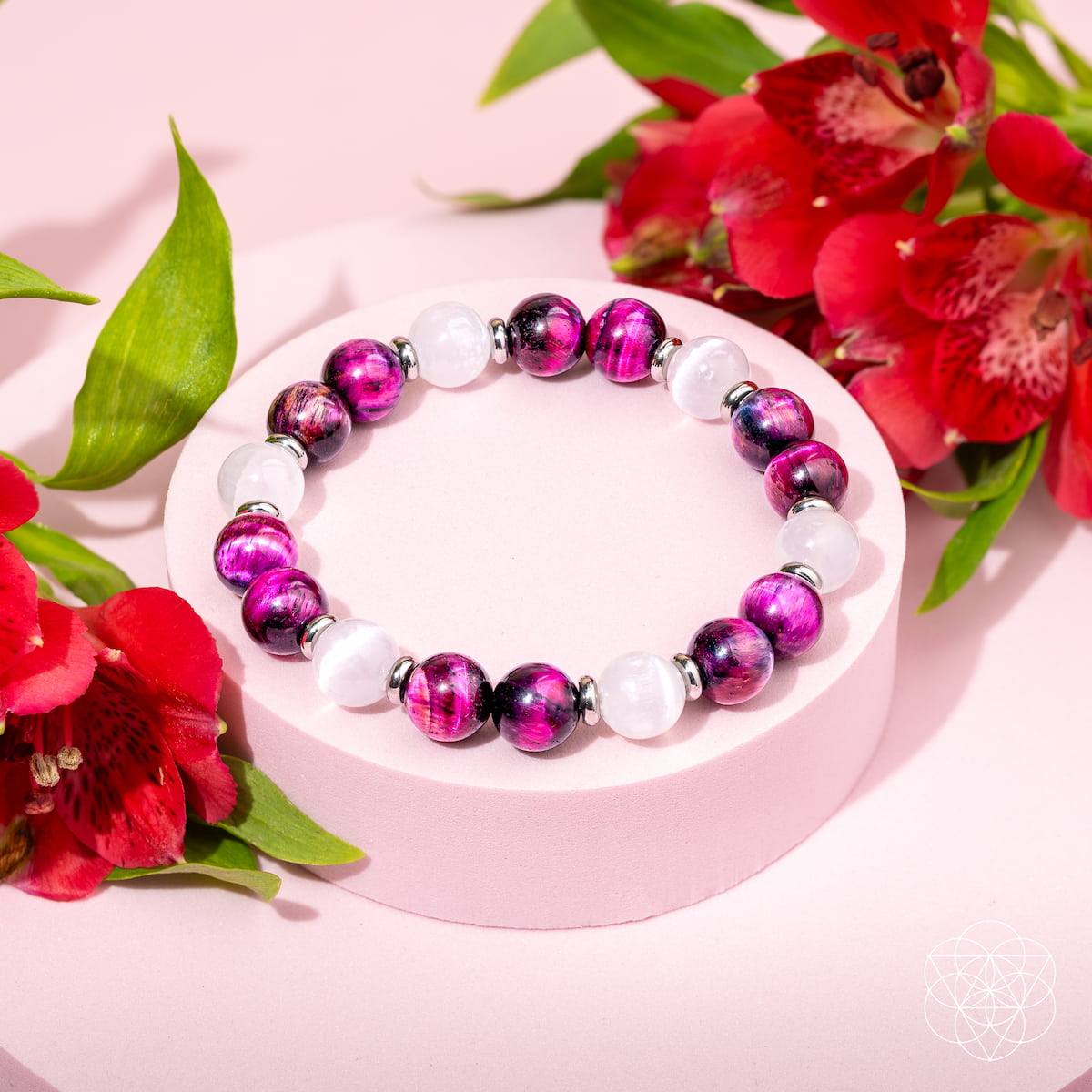 The Shape Your Future – Pink Galaxy Tiger’s Eye Bracelet by Conscious Items, featuring alternating purple and white beads, is displayed on a round pink platform with red flowers and green leaves set against a pink background.