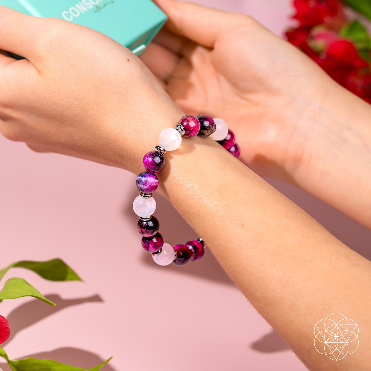 Wearing the Shape Your Future – Pink Galaxy Tiger’s Eye Bracelet by Conscious Items, featuring pink, purple, and white stones, a person holds a turquoise box. The pink background has red flowers and green leaves; a white geometric logo is bottom right.