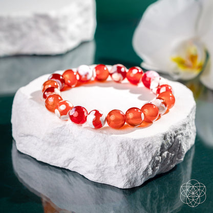 The Feminine Force – Carnelian Orca Agate Bracelet by Conscious Items, featuring shiny red and white marbled beads, is showcased on a textured white stone with a blurred white flower and soft green background.