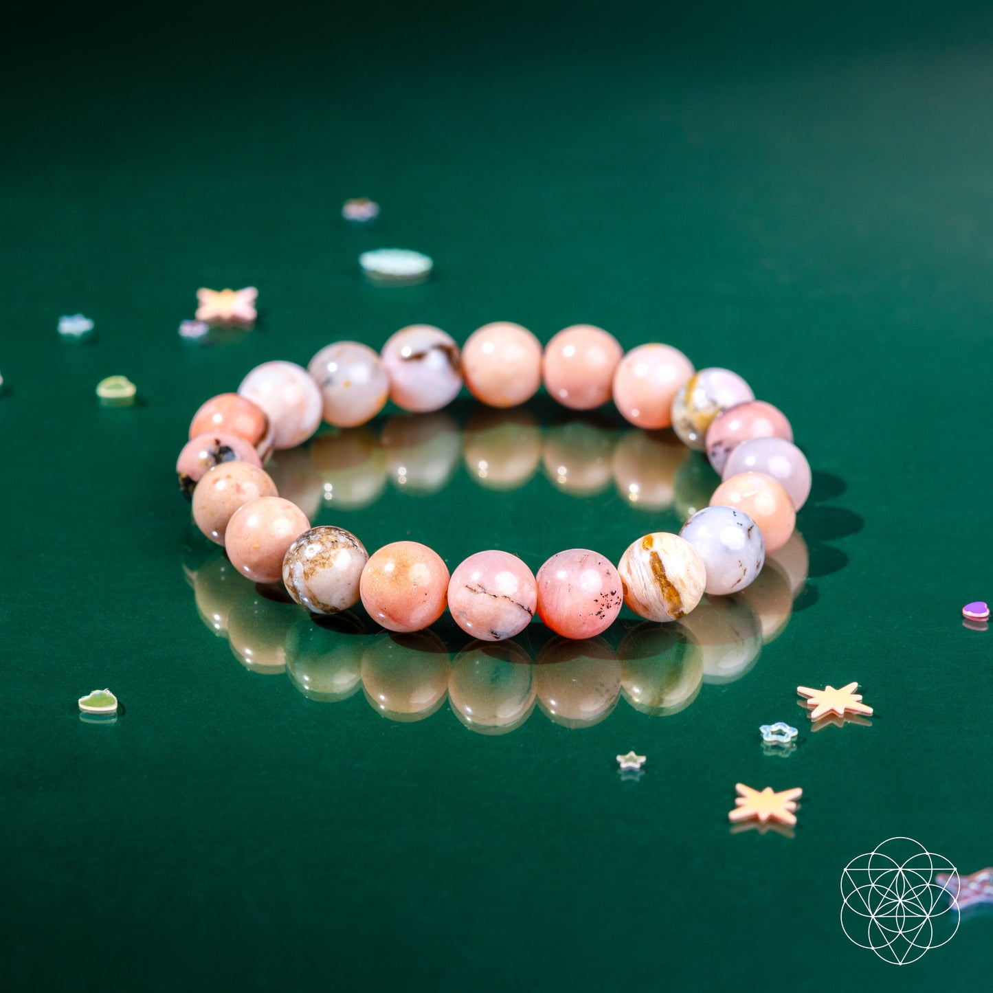 The Repair My Heart - The Peruvian Pink Opal Bracelet by Conscious Items, featuring pink, lavender, and marbled beads for heart healing, rests on a glossy green surface amid star and heart decorations. A geometric white logo appears at the bottom right.