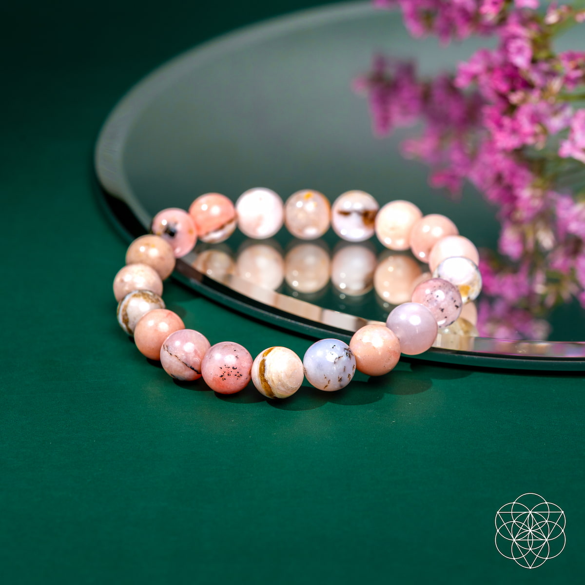 The Repair My Heart - The Peruvian Pink Opal Bracelet by Conscious Items, a pink and white beaded piece for heart healing and emotional release, rests on a round mirror with pink flower reflections on green, featuring a geometric white logo.