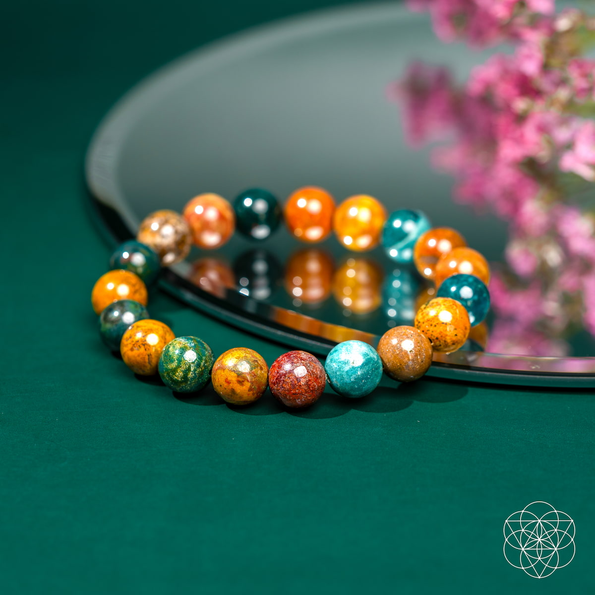 The Quiet My Mind – Ocean Jasper Bracelet by Conscious Items features vibrant beads in orange, green, blue, and brown, displayed on a green surface beside a mirror with pink flowers, symbolizing emotional balance.