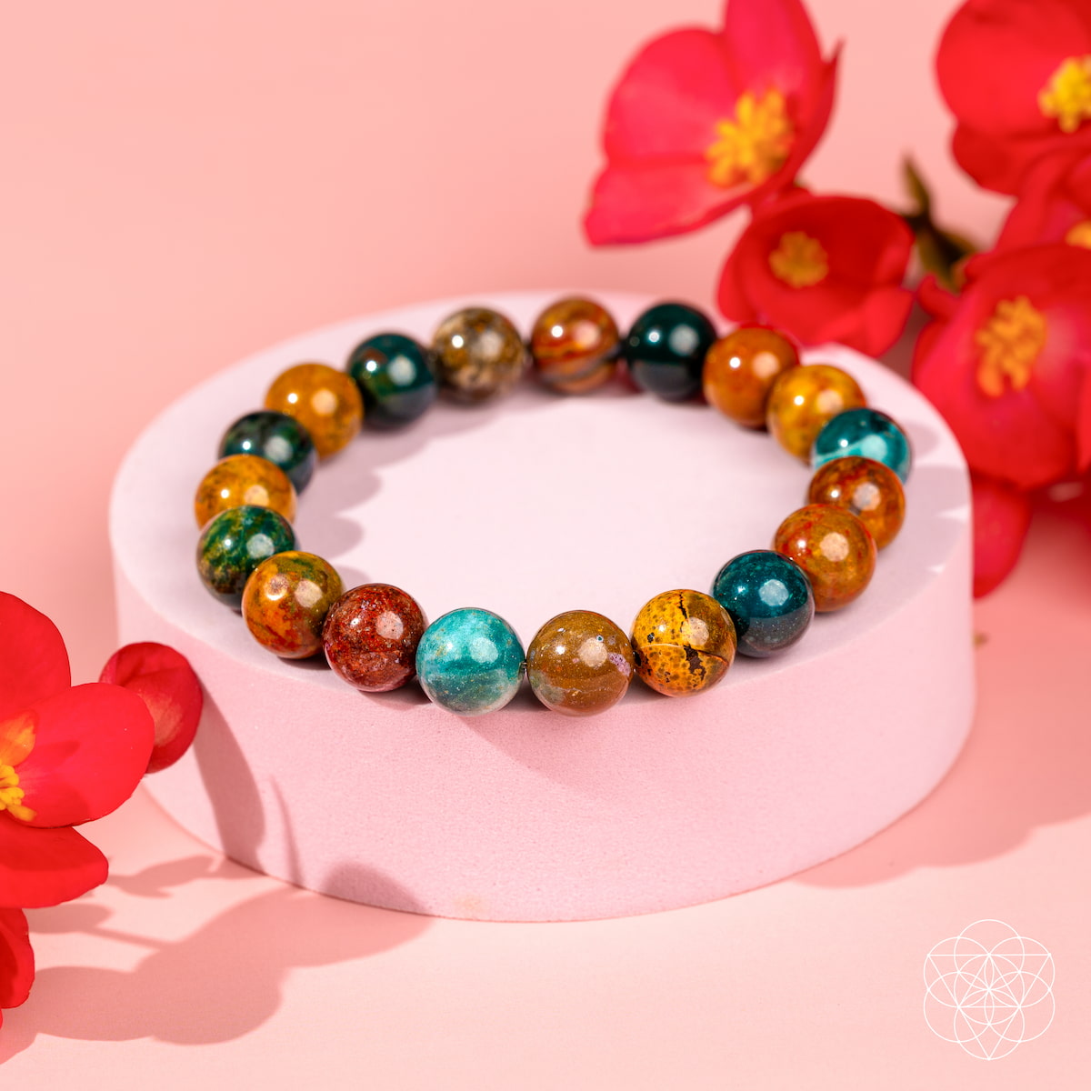 The Quiet My Mind – Ocean Jasper Bracelet by Conscious Items, featuring green, red, yellow, and ocean jasper beads, rests on a round pink platform with vibrant red flowers against a soft pink background for emotional balance.
