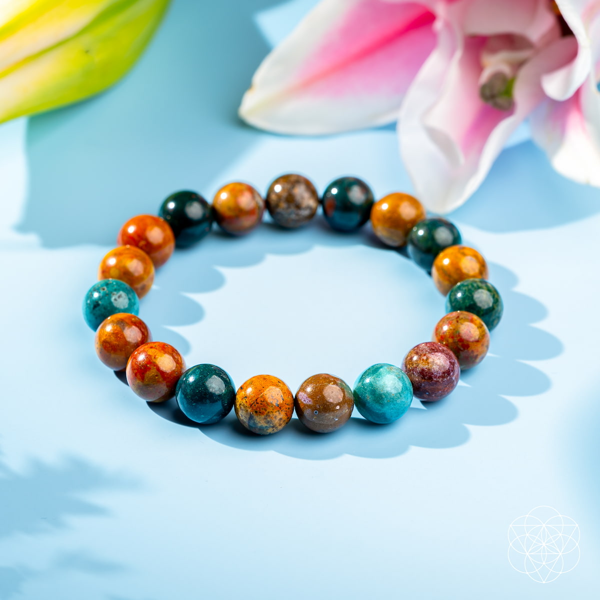 The Quiet My Mind – Ocean Jasper Bracelet by Conscious Items is displayed on a light blue surface next to a pink lily, with soft lighting creating gentle shadows and an elegant, calming atmosphere to enhance emotional balance.
