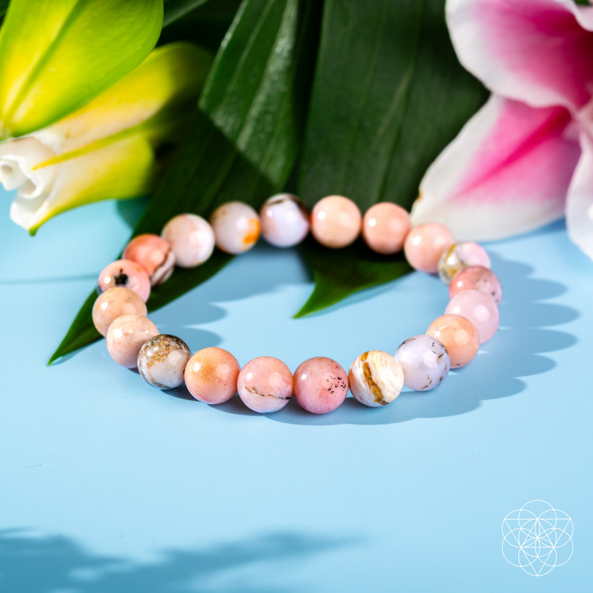 The Repair My Heart - The Peruvian Pink Opal Bracelet by Conscious Items, featuring pink, white, and brown marbled stones for heart healing, is displayed on a light blue surface with green leaves and vibrant yellow and pink flowers.