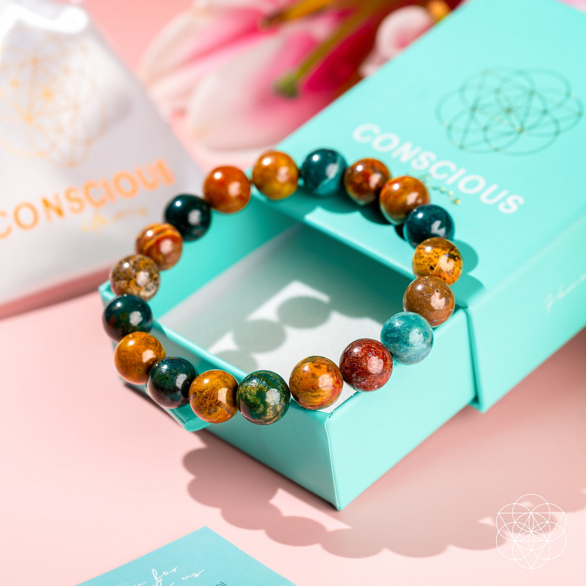 The Quiet My Mind – Ocean Jasper Bracelet by Conscious Items is displayed on an open turquoise branded box with a white pouch and pink flower on a pastel pink surface—designed to support emotional balance and anxiety relief.