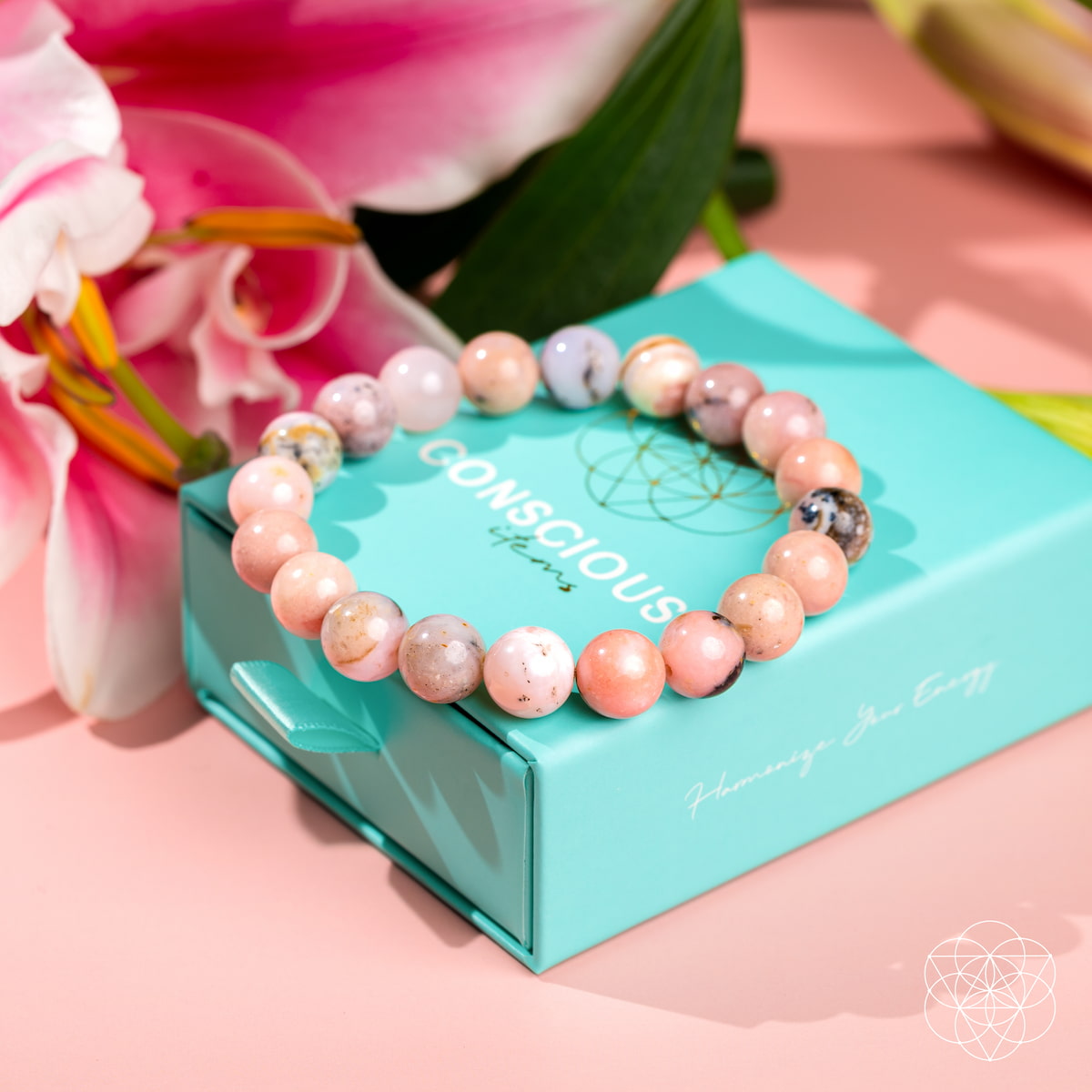 The Repair My Heart - The Peruvian Pink Opal Bracelet by Conscious Items, adorned with pink and beige beads for heart healing and emotional release, sits on a turquoise branded box near pink lilies atop a pastel pink surface. White geometric logo bottom right.