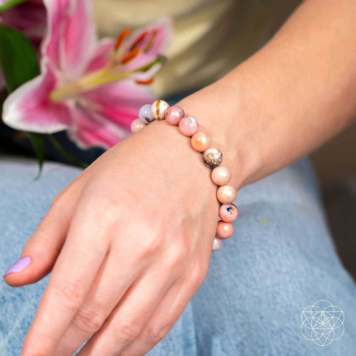 A hand with light nail polish wears the "Repair My Heart - The Peruvian Pink Opal Bracelet" by Conscious Items, resting on blue jeans and holding a pink lily. The pastel beads support heart healing and emotional release; the background is softly blurred.
