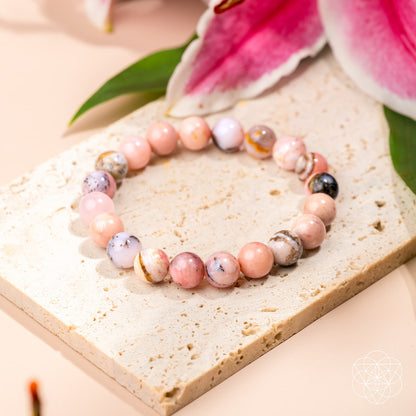 The Repair My Heart - The Peruvian Pink Opal Bracelet by Conscious Items, featuring round pink, white, and brown marbled stones for heart healing, is displayed on a beige tile with pink lilies and green leaves in the background.