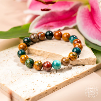 The Quiet My Mind – Ocean Jasper Bracelet by Conscious Items features polished Ocean Jasper beads, displayed on beige stone tiles with pink lilies and green leaves—perfect for emotional balance and subtle anxiety relief.