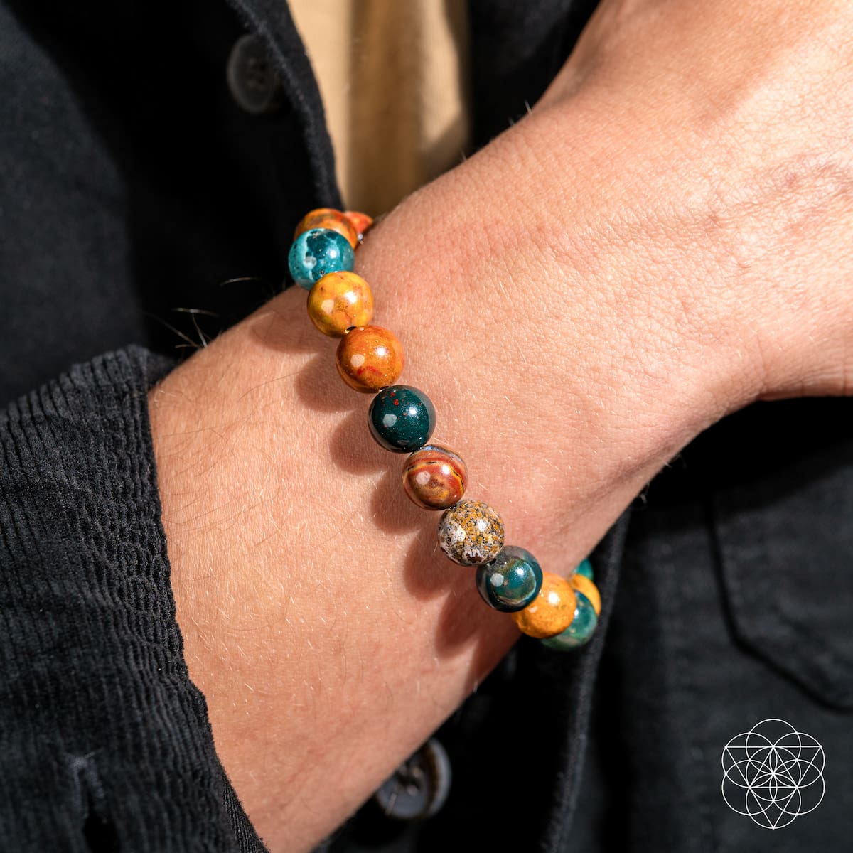 A close-up of a wrist adorned with the Quiet My Mind – Ocean Jasper Bracelet by Conscious Items, featuring round green, yellow, and orange stones. The wearer sports a black textured jacket, and a geometric logo is visible in the corner.