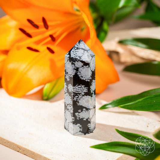 The Spiritual Bodyguard - Snowflake Obsidian Point by Conscious Items, celebrated for emotional balance and stability, stands on a light stone surface with an orange lily and green leaves. A white geometric logo appears in the bottom right corner.