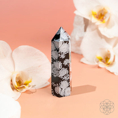 The Spiritual Bodyguard - Snowflake Obsidian Point by Conscious Items stands upright on a peach background, framed by white orchids with yellow centers—a refined symbol of balance and stability.