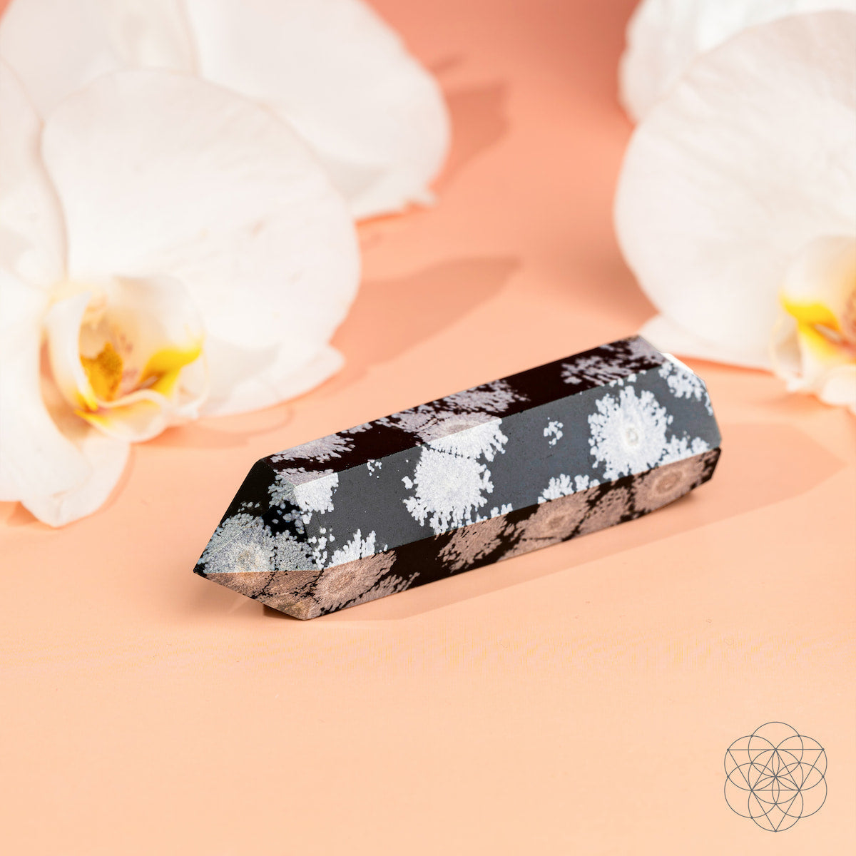 The Conscious Items Spiritual Bodyguard - Snowflake Obsidian Point, a polished black crystal with white splotches, rests on a peach surface between two white orchids, symbolizing balance and stability.