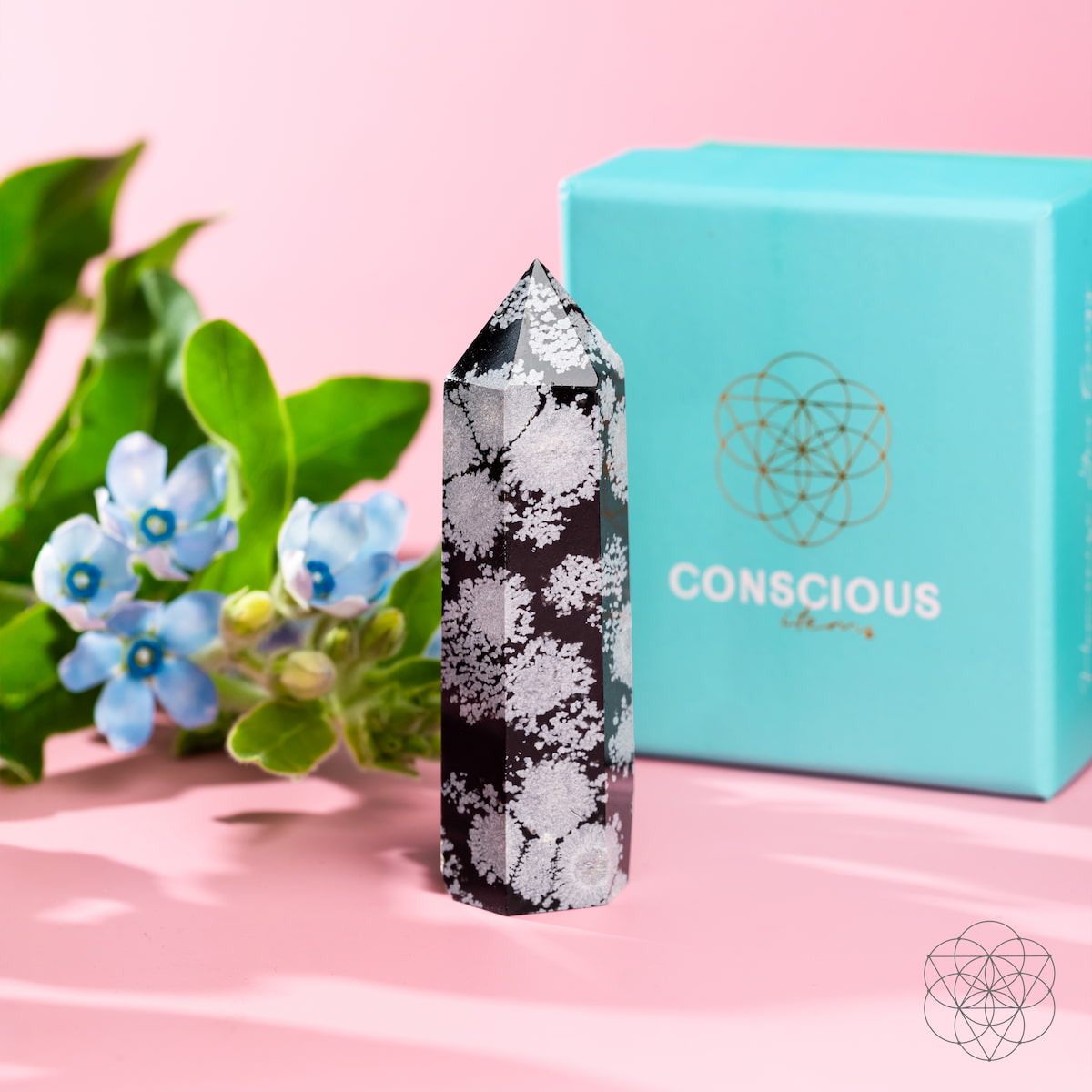 The Spiritual Bodyguard - Snowflake Obsidian Point by Conscious Items, featuring white snowflake-like patterns, is displayed beside blue flowers and green leaves with a turquoise Conscious Items box on a pink surface.
