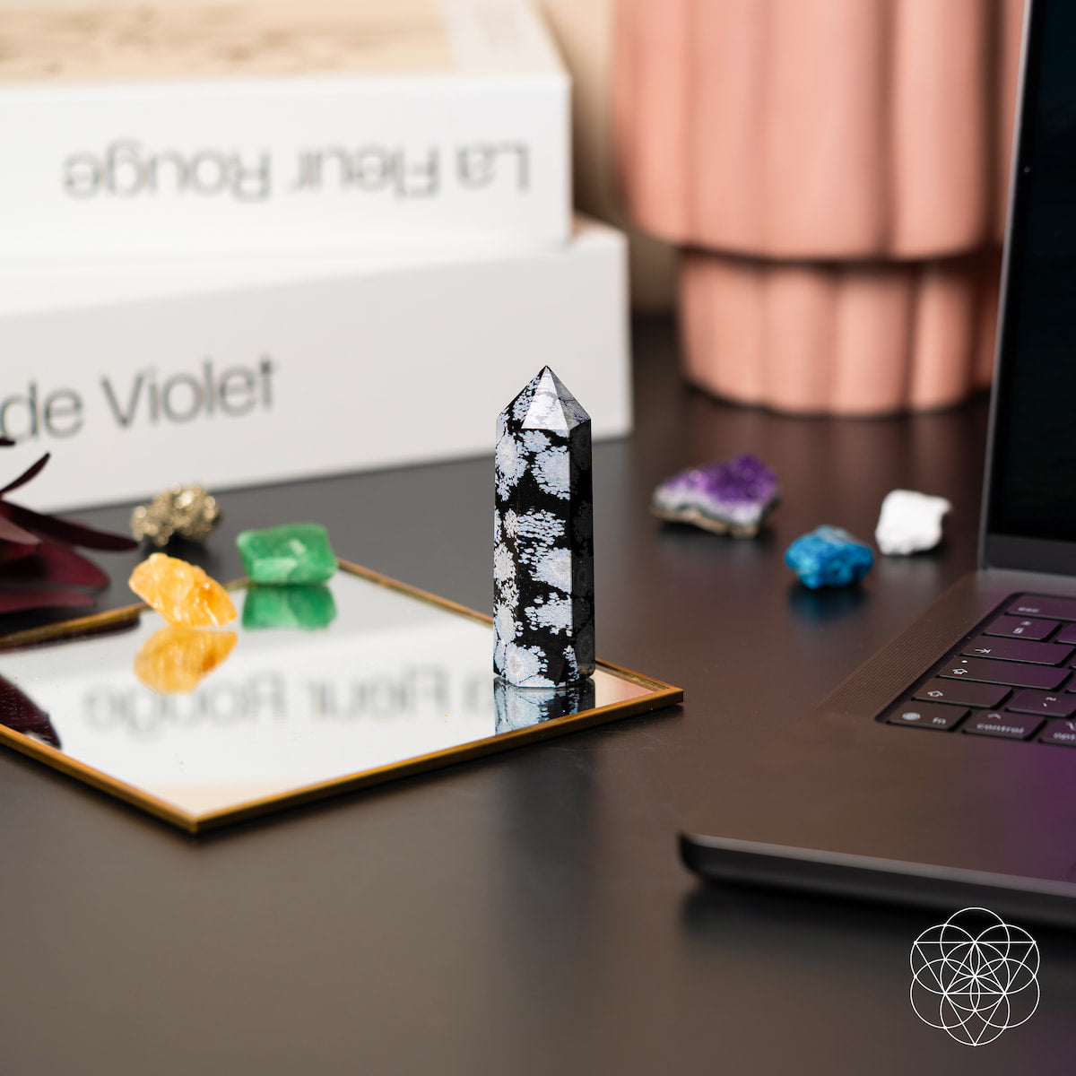 The Spiritual Bodyguard - Snowflake Obsidian Point by Conscious Items, known for balance and stability, stands on a small mirror next to colorful crystals, a laptop, and stacked books on a stylish black desk.