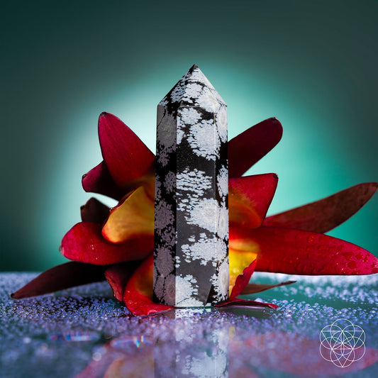 The Spiritual Bodyguard - Snowflake Obsidian Point by Conscious Items, an emotional healing stone for balance and stability, stands upright before a red and yellow flower with water droplets on a reflective surface against a green-blue gradient background.