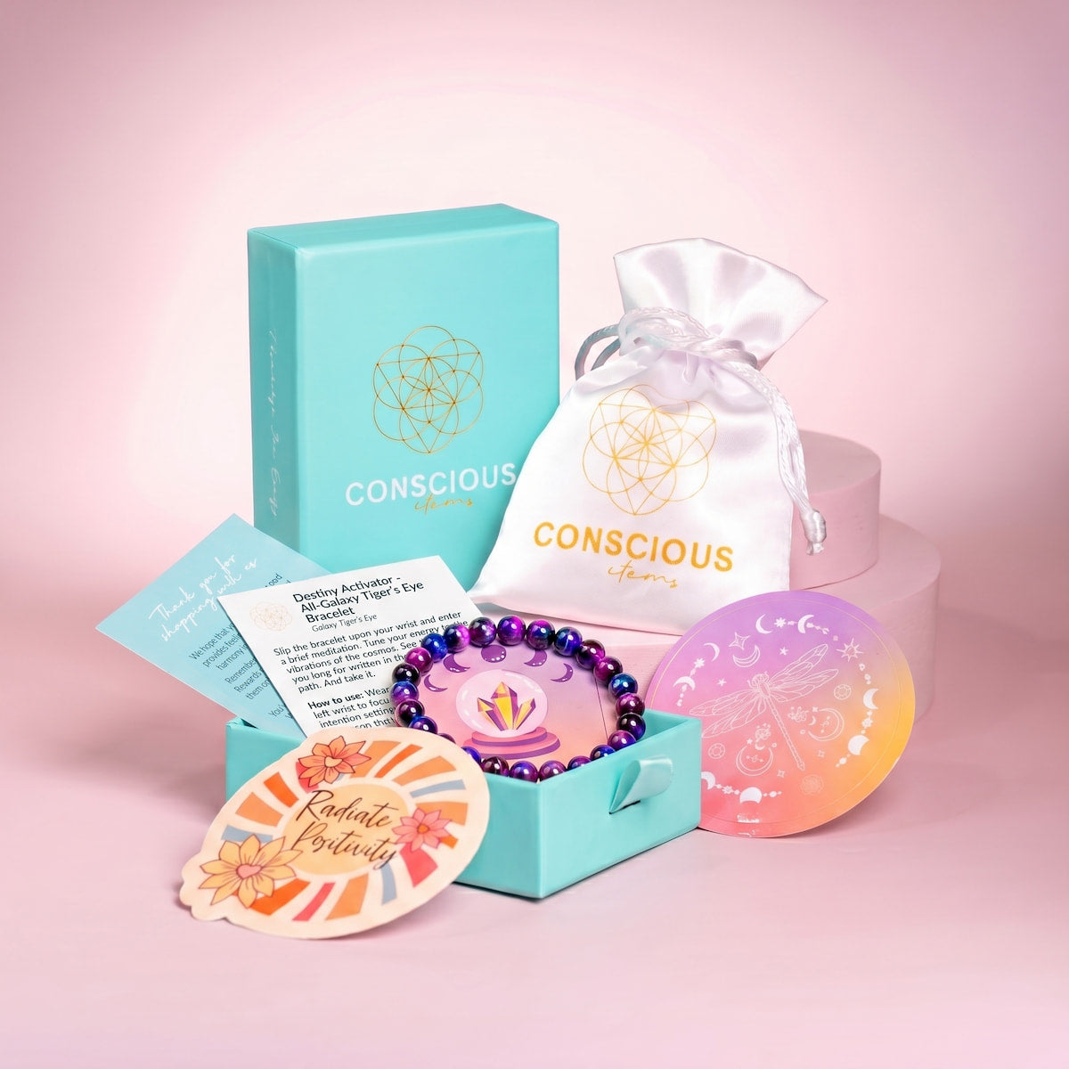 The Destiny Activator - All-Galaxy Tiger’s Eye Bracelet by Conscious Items comes in a teal gift box with a drawstring pouch, info card, and illustrated cards—beautifully displayed on pastel pink platforms to help you embrace your cosmic destiny.