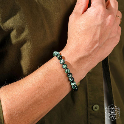 A close-up of a wrist adorned with the Angel Energy – Rare Seraphinite Bracelet of Transformation by Conscious Items. Paired with an olive-green shirt, the wearer’s relaxed hand highlights this transformation stone’s natural beauty.