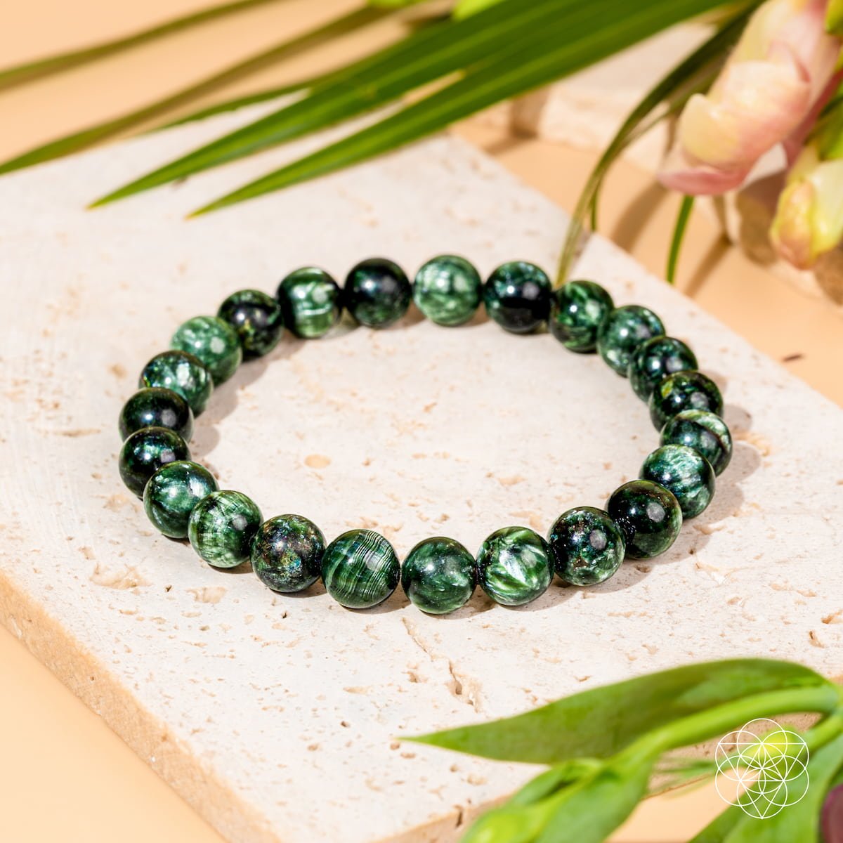 The Angel Energy – Rare Seraphinite Bracelet of Transformation by Conscious Items, crafted from polished green seraphinite stones, rests on a textured white tile with green leaves and pink flowers, symbolizing renewal and transformation.