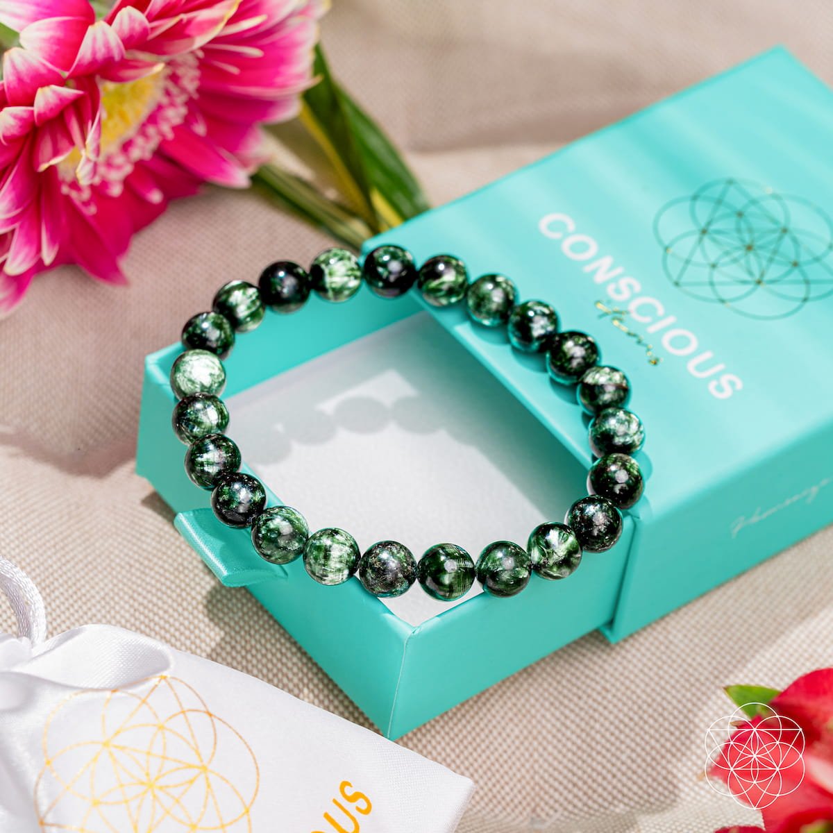 The Angel Energy – Rare Seraphinite Bracelet of Transformation by Conscious Items sits atop a turquoise box, surrounded by pink flowers and a white pouch on beige fabric—a beautiful display for gifting this transformation stone.