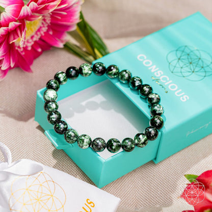 The Angel Energy – Rare Seraphinite Bracelet of Transformation by Conscious Items sits atop a turquoise box, surrounded by pink flowers and a white pouch on beige fabric—a beautiful display for gifting this transformation stone.