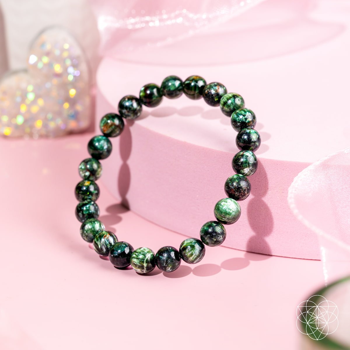 The Angel Energy – Rare Seraphinite Bracelet of Transformation by Conscious Items shines on a round pink platform with a sparkly heart in the background and a soft pink ribbon, highlighting this stunning transformation stone.