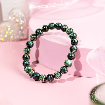 The Angel Energy – Rare Seraphinite Bracelet of Transformation by Conscious Items shines on a round pink platform with a sparkly heart in the background and a soft pink ribbon, highlighting this stunning transformation stone.