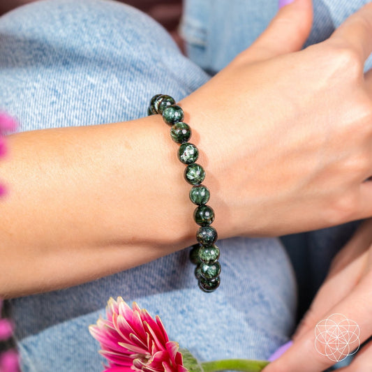 A person wearing light blue jeans showcases the Conscious Items Angel Energy – Rare Seraphinite Bracelet of Transformation on their wrist, with light purple nails and pink flowers in the foreground.
