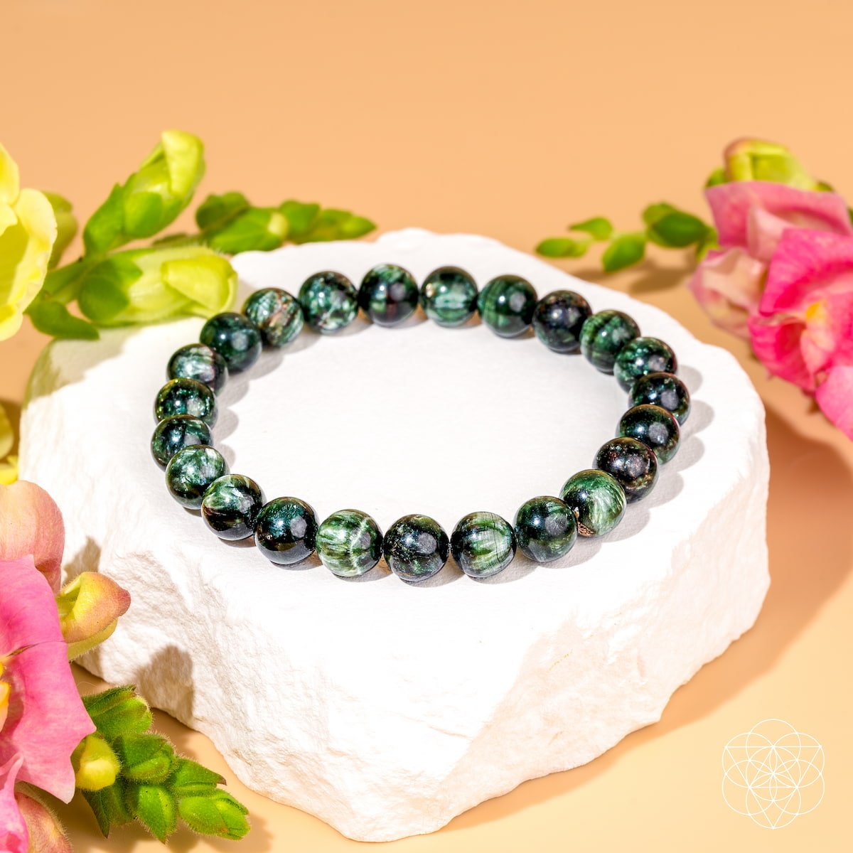The Angel Energy – Rare Seraphinite Bracelet of Transformation by Conscious Items, featuring round green and black beads, is displayed on a white stone platform with pink and yellow flowers against a soft peach background.
