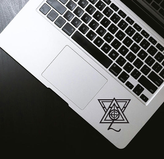 A silver laptop on a black surface with a geometric black sticker featuring overlapping triangles and a circle on the bottom right corner of the palm rest.