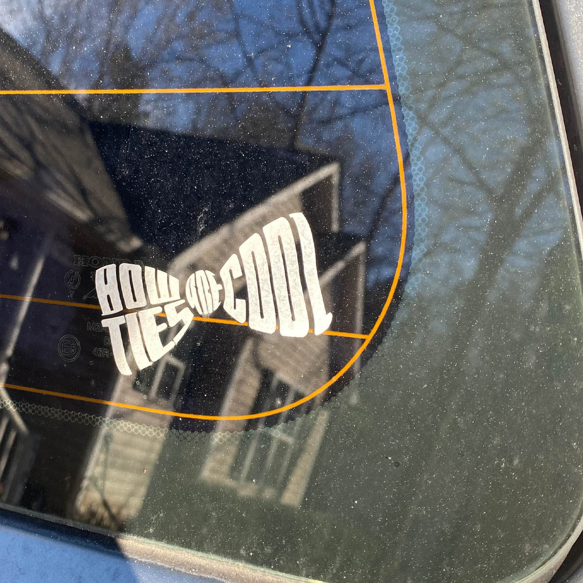 Car window with a 'Bowties are Cool' sticker reflecting a house and trees.