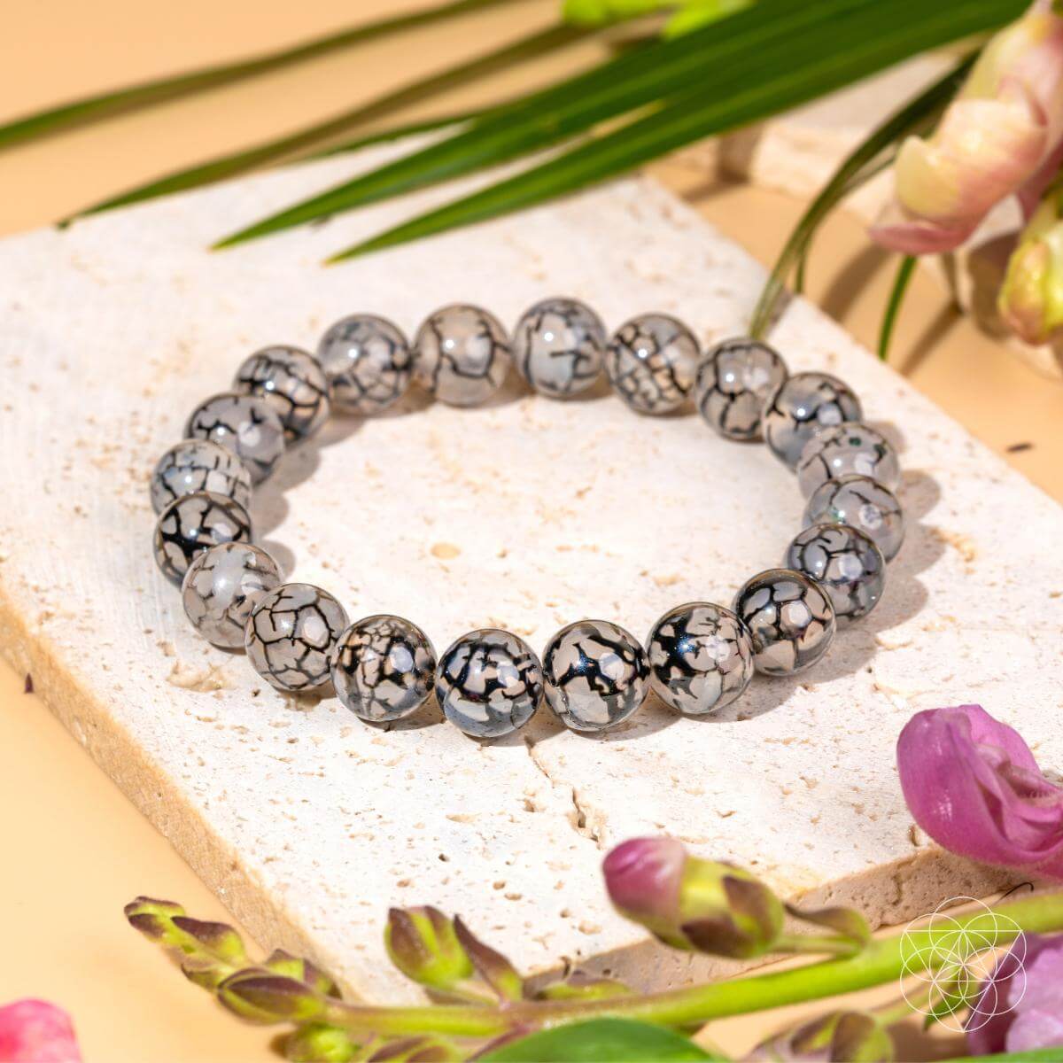 The Calm in the Chaos - River Agate Bracelet by Conscious Items features round, marbled black and gray beads. Displayed on a stone slab with green leaves and pink flowers, this calming bracelet is known for stress relief.