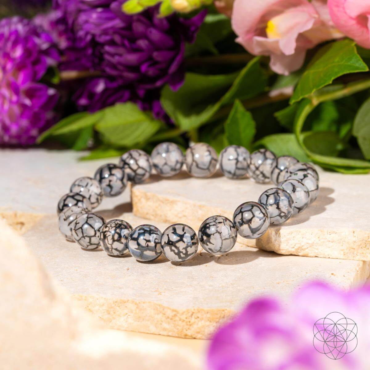 The Calm in the Chaos - River Agate Bracelet by Conscious Items features gray and black beads, shown on light stone tiles among vibrant purple and pink flowers, offering natural stress relief.
