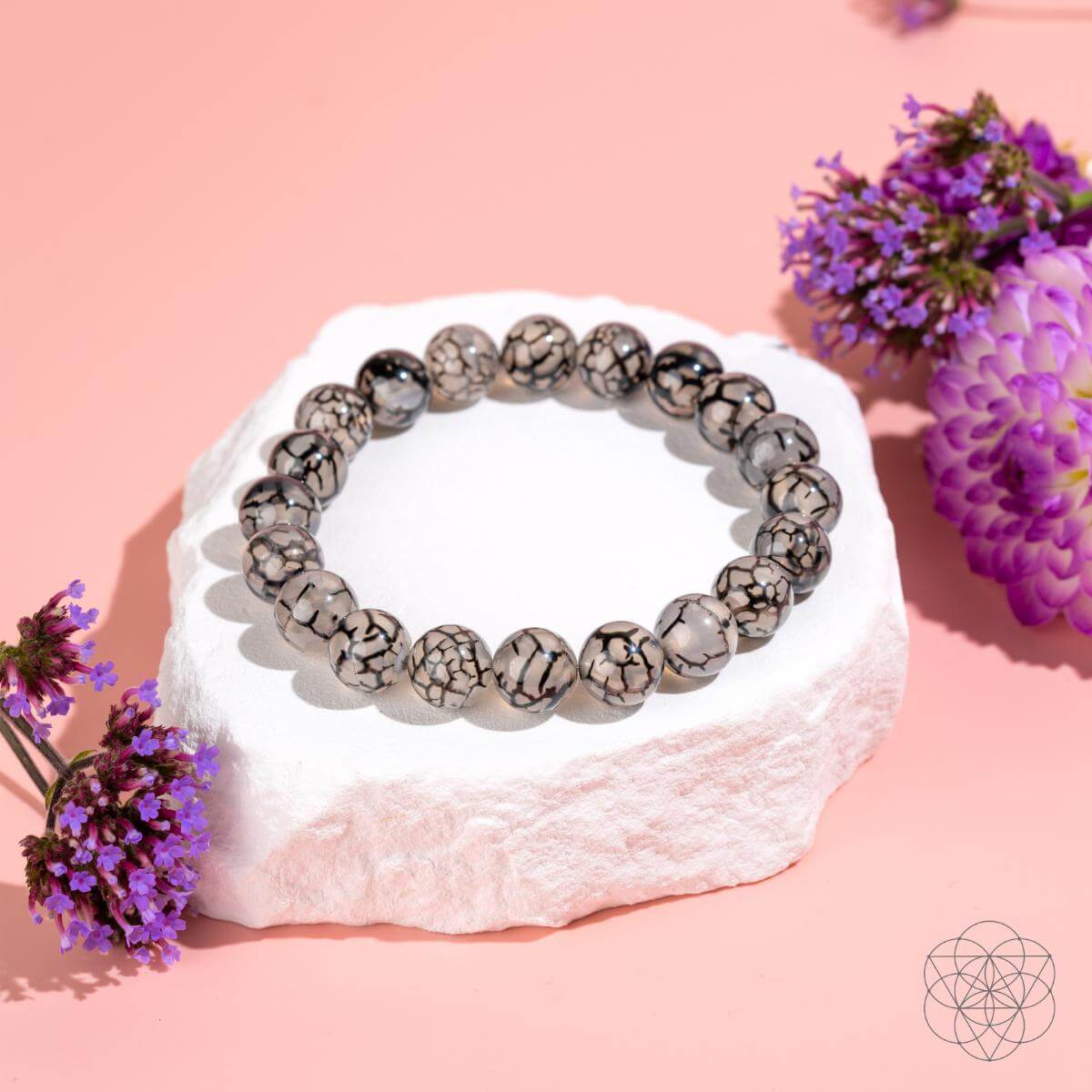 The Calm in the Chaos - River Agate Bracelet by Conscious Items, featuring black and white marbled agate stones, is displayed on a white textured surface with small purple flowers on a soft pink background.