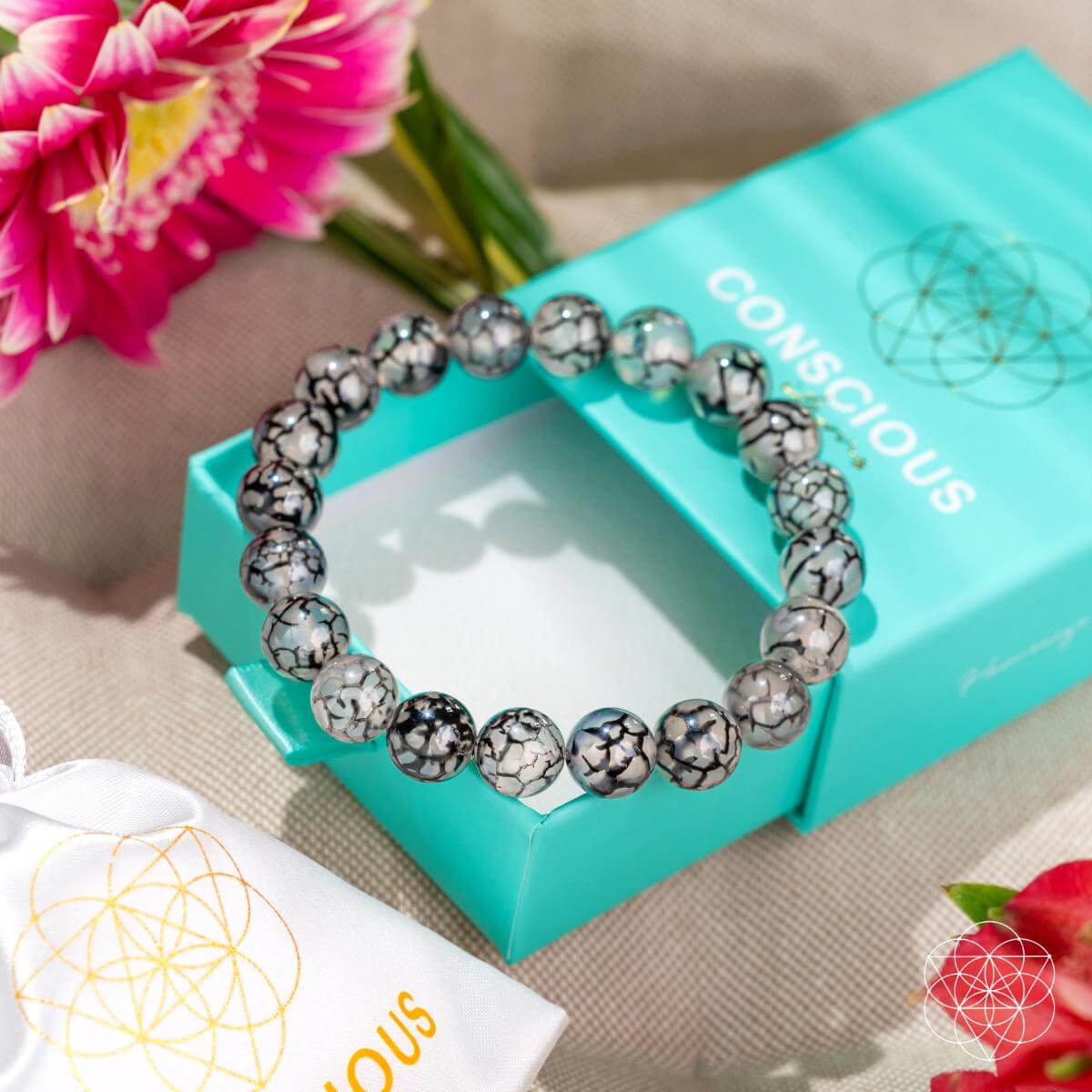 The Calm in the Chaos - River Agate Bracelet by Conscious Items, a stress-relieving black and white marble bead accessory, is displayed atop a teal CONSCIOUS box beside pink flowers and a white pouch with a gold geometric pattern.