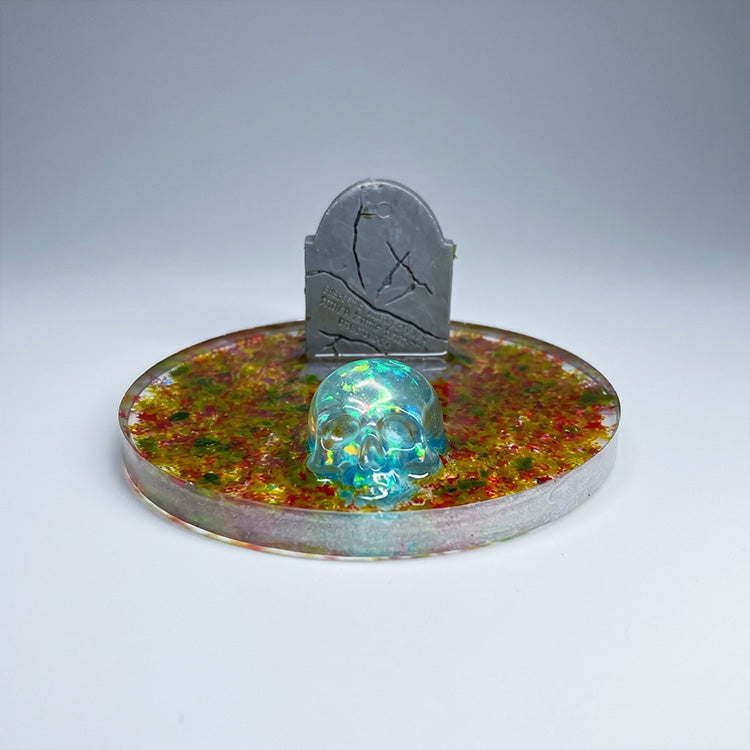 Glowing blue skull on a colorful circular base with a gray tombstone against a gray background