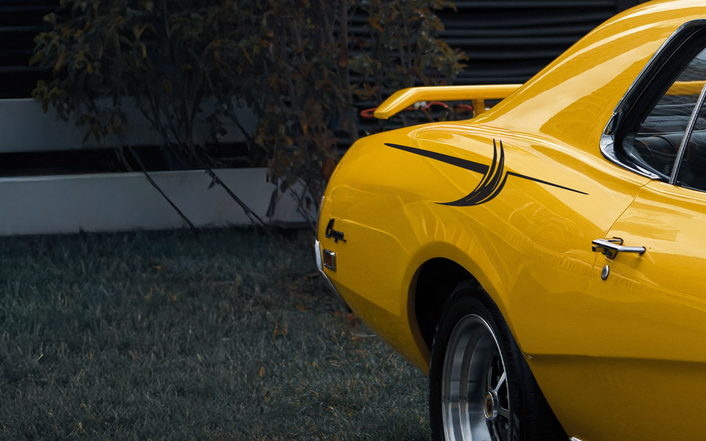 The rear side of a bright yellow vintage muscle car with a black stripe detail and a rear spoiler, parked on grass near bushes and a modern building.