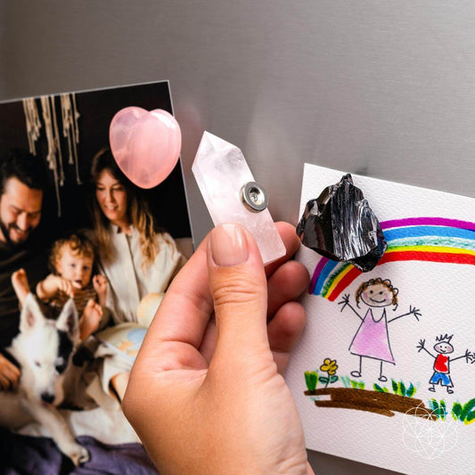 A hand attaches a Conscious Items Crystal Guardian Magnet—part of the Trio of Peace & Protection—to the fridge, securing family mementos. Nearby are the heart-shaped Rose Quartz and dark Black Obsidian magnets from the same set.