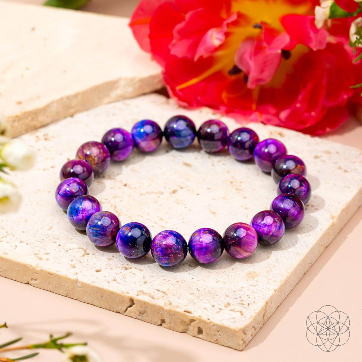 The Destiny Activator - All-Galaxy Tiger’s Eye Bracelet by Conscious Items, featuring glossy purple and blue marbled beads, rests on a cream stone surface among pink and red flowers. A geometric logo appears in the bottom right corner.